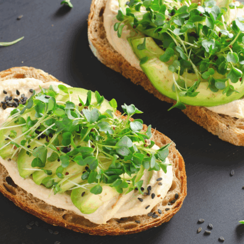 A slice of rustic sourdough toast generously topped with creamy hummus and fresh avocado slices, sprinkled with microgreens and a hint of chilli flakes, served on a ceramic plate—a vibrant, nutritious breakfast or snack.