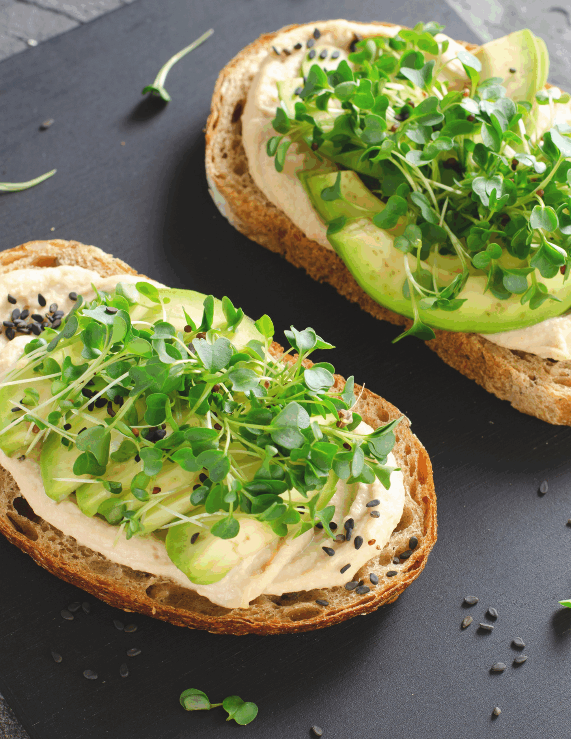 A slice of rustic sourdough toast generously topped with creamy hummus and fresh avocado slices, sprinkled with microgreens and a hint of chilli flakes, served on a ceramic plate—a vibrant, nutritious breakfast or snack.
