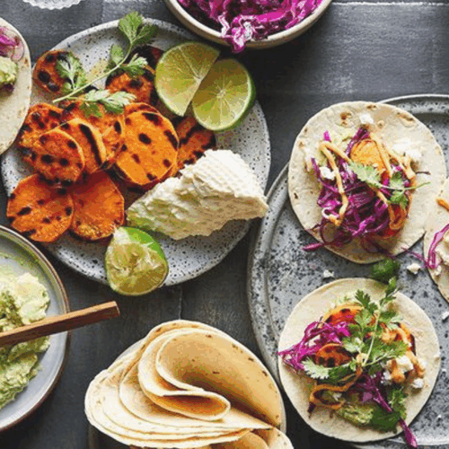 A colourful plate of grilled sweet potato tacos, topped with fresh avocado, tangy feta, crisp red cabbage, and a sprinkle of cilantro, served in warm corn tortillas with a wedge of lime.
