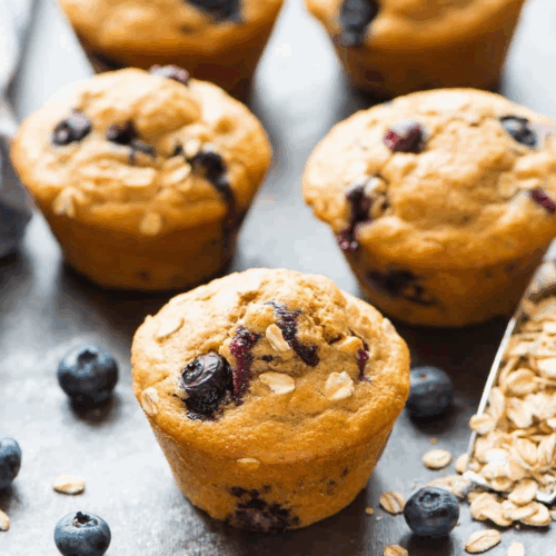 Golden Healthy Blueberry Muffins bursting with fresh blueberries, stacked on a white plate and garnished with a dusting of oats.