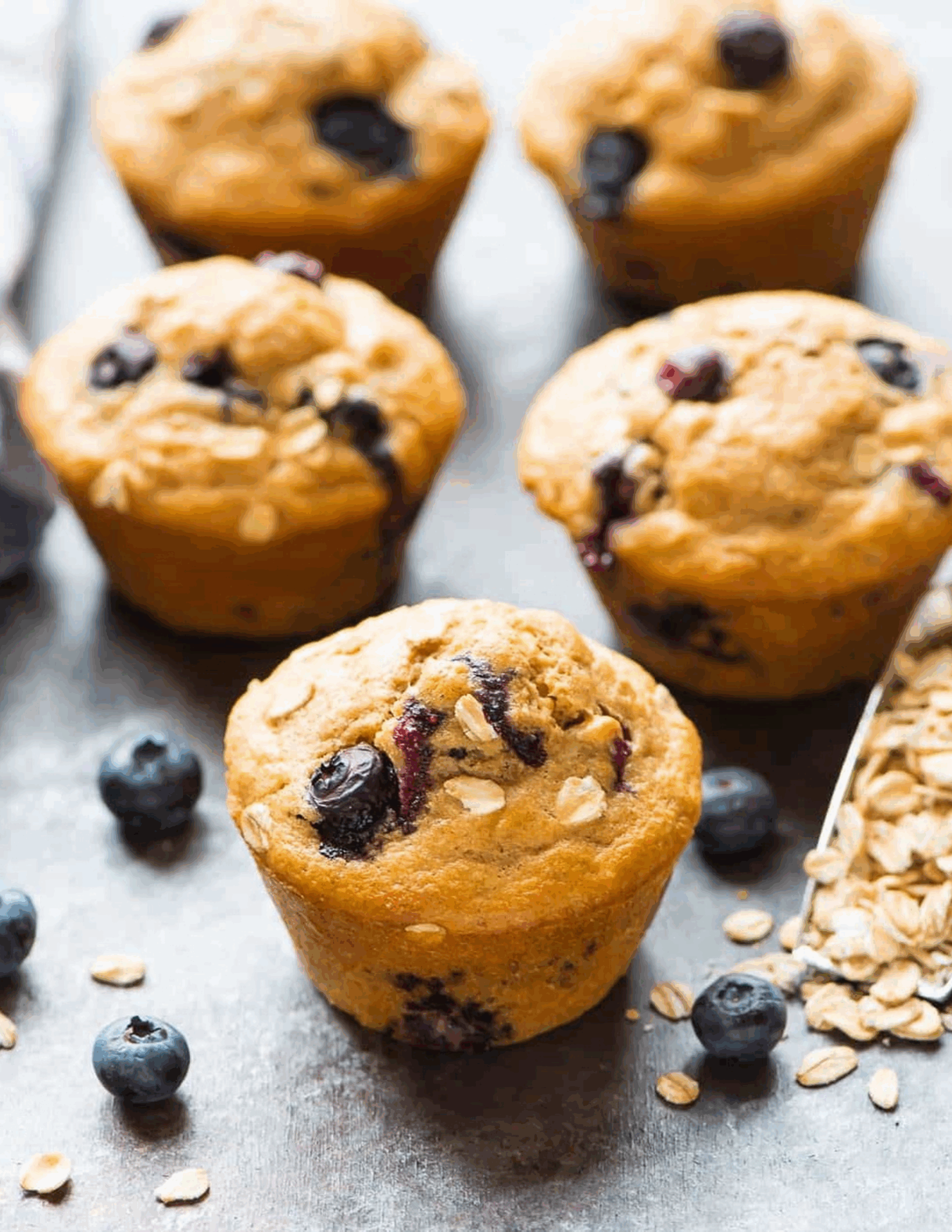 Golden Healthy Blueberry Muffins bursting with fresh blueberries, stacked on a white plate and garnished with a dusting of oats.