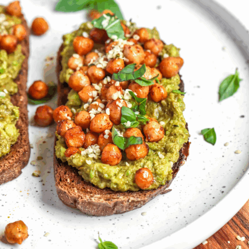 A rustic slice of wholegrain toast topped with creamy pesto chickpea mash, juicy cherry tomatoes, and a sprinkle of fresh basil, served on a ceramic plate for a vibrant, nutritious breakfast.