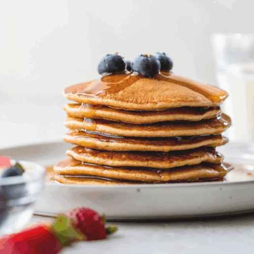 A stack of fluffy Protein Pancakes topped with fresh berries and a drizzle of maple syrup, served on a rustic ceramic plate.