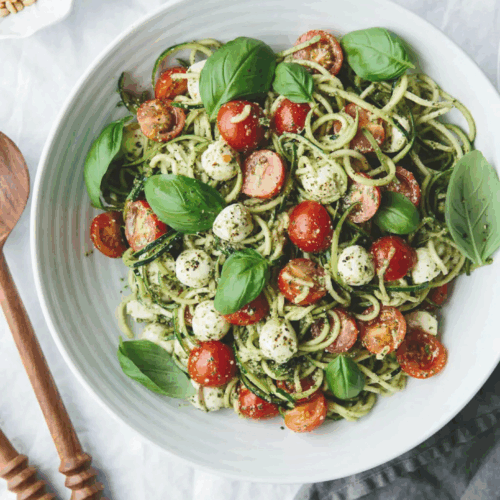 Spiralized zucchini noodles tossed with juicy cherry tomatoes, creamy mozzarella balls, and fresh basil leaves, drizzled with balsamic glaze and arranged on a bright ceramic plate.
