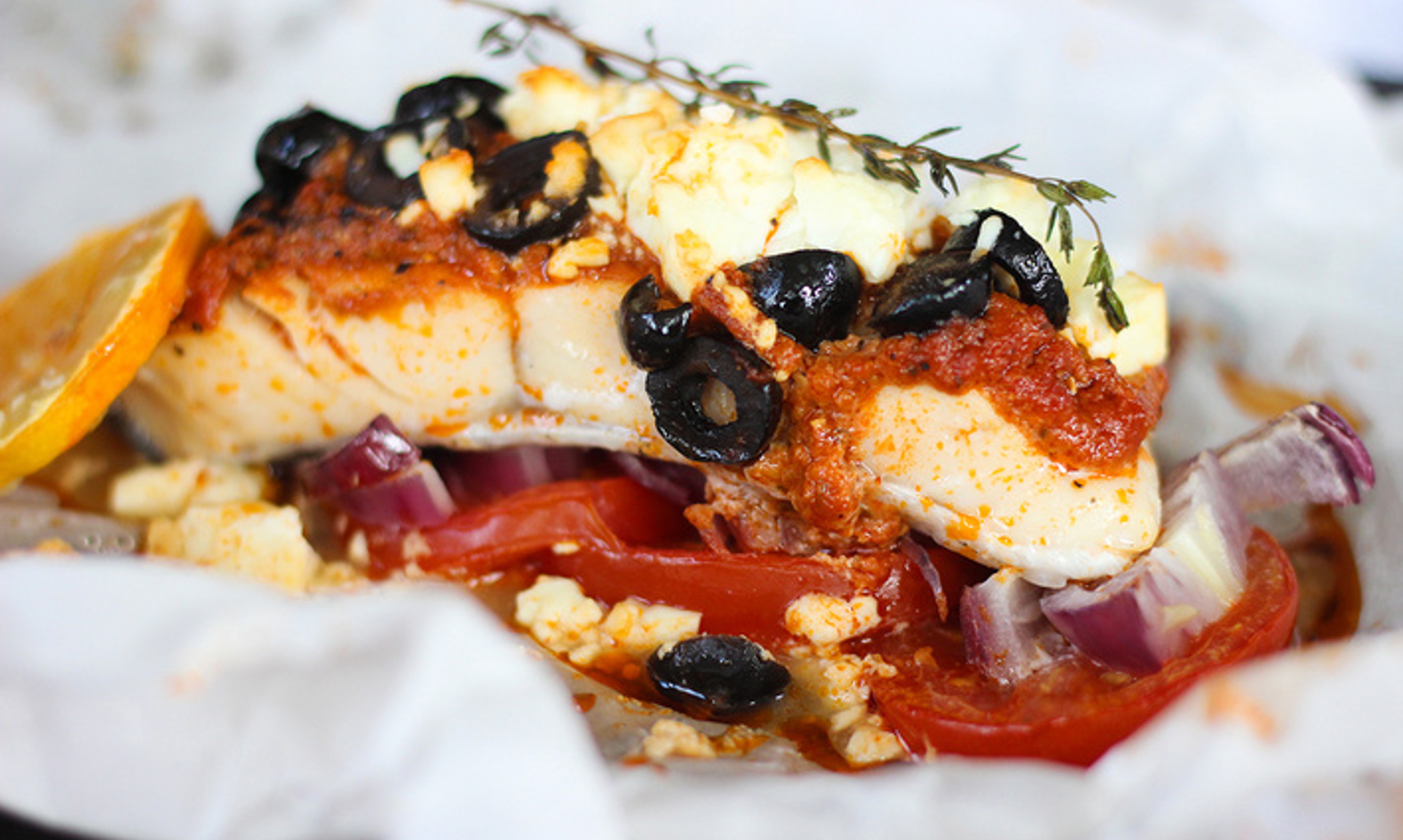 Baked white fish fillet topped with cherry tomatoes, black olives, herbs, and a drizzle of olive oil on a ceramic dish