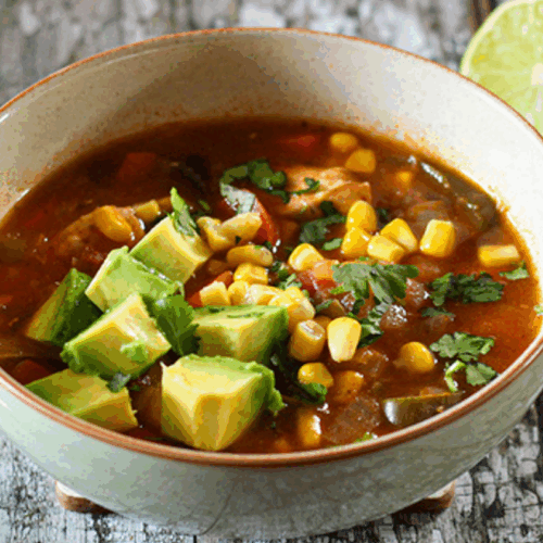 Bowl of Mexican chicken soup with shredded chicken, corn, beans, tomatoes, and herbs garnished with lime and avocado