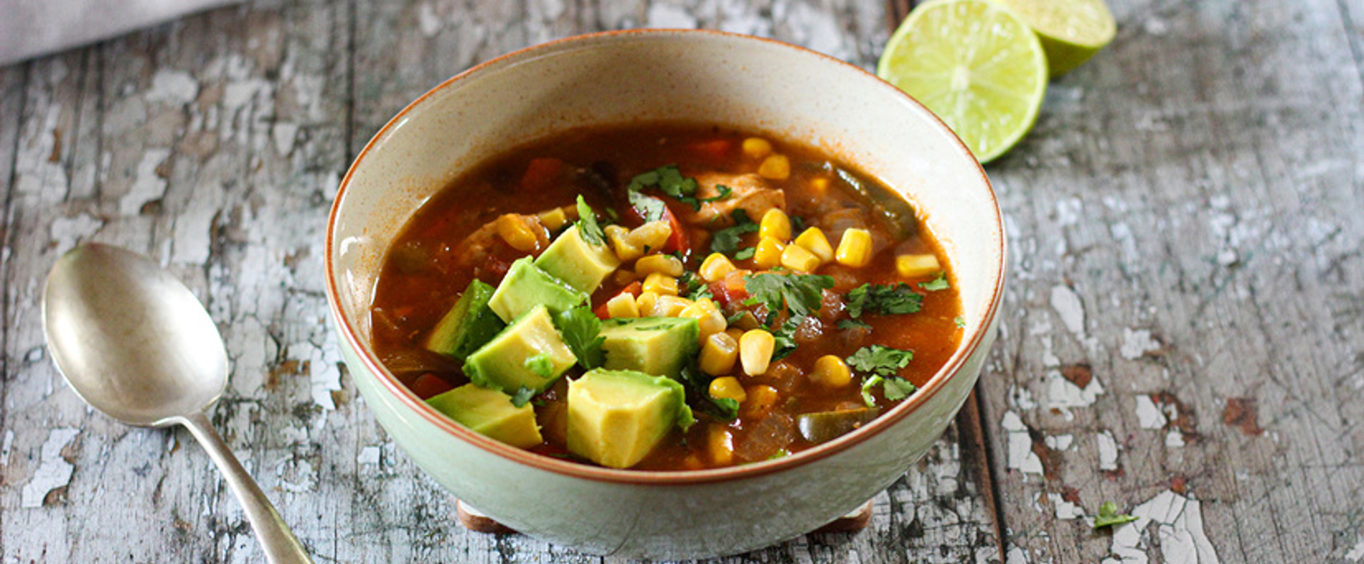 Bowl of Mexican chicken soup with shredded chicken, corn, beans, tomatoes, and herbs garnished with lime and avocado